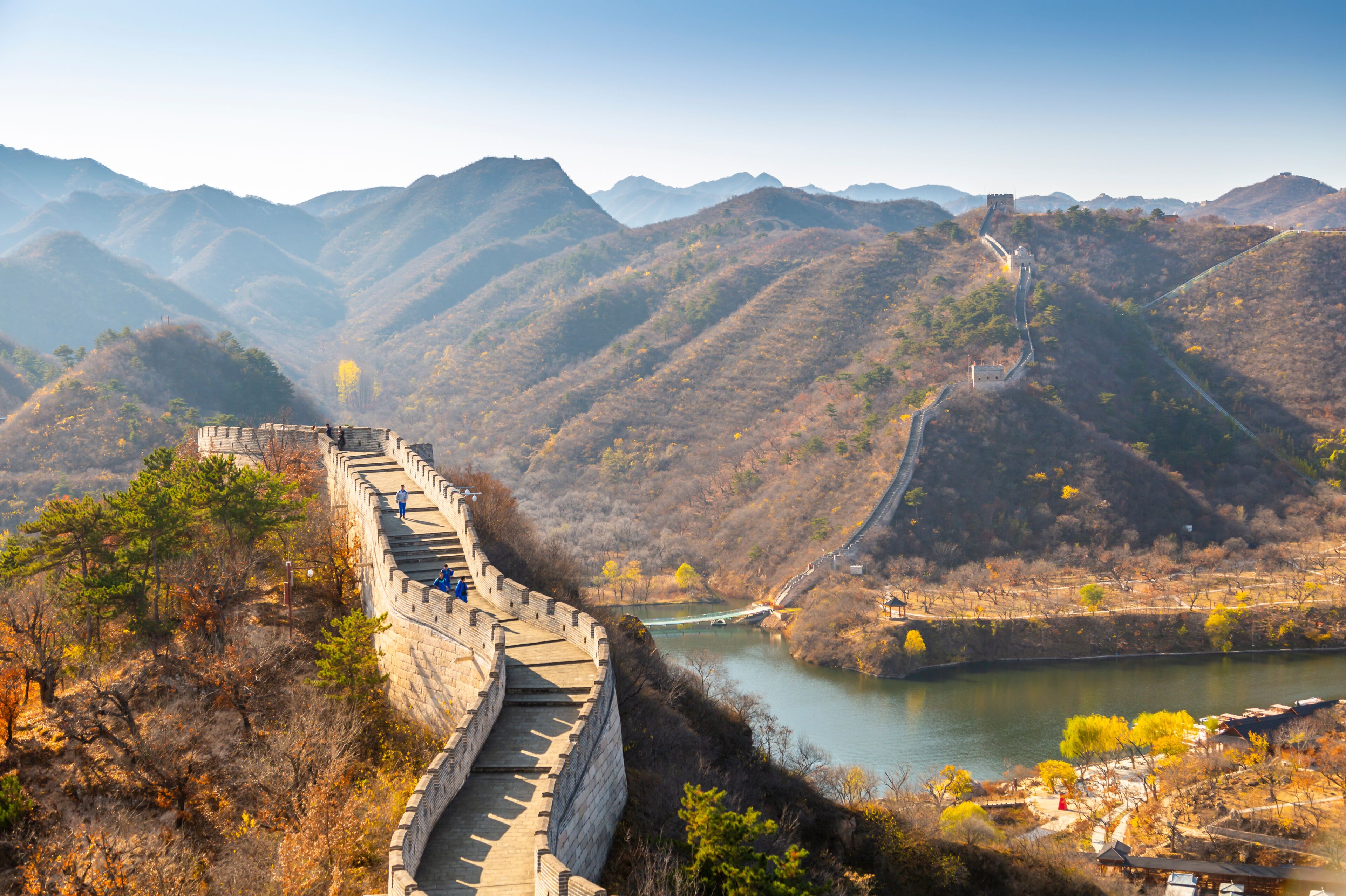 The Great Wall of China winding through a mountainous landscape with a river and traditional Chinese architecture.