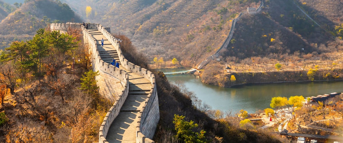 The Great Wall of China winding through a mountainous landscape with a river and traditional Chinese architecture.