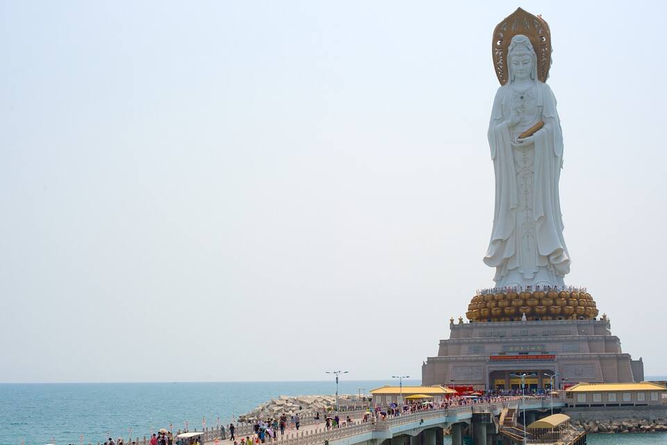 A large white statue of a deity with a golden base, located near a body of water with a viewing platform.
