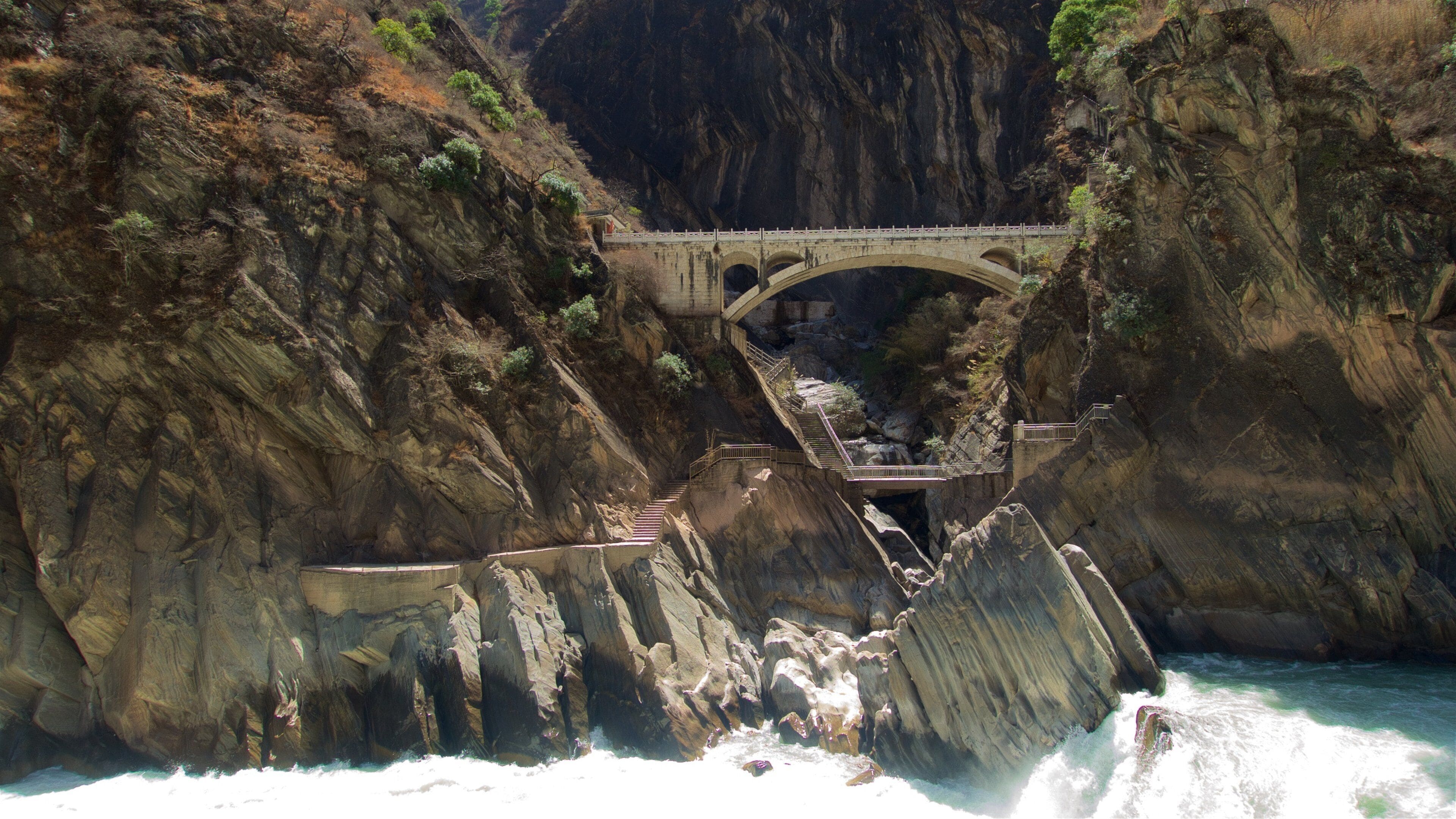 Un puente de piedra sobre un río en un cañón, con acantilados rocosos y escaleras que bajan hacia el agua.