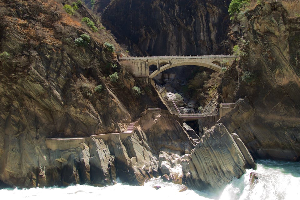 Un puente de piedra sobre un río en un cañón, con acantilados rocosos y escaleras que bajan hacia el agua.