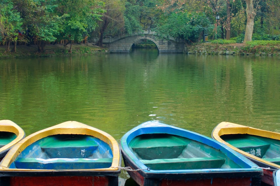 A row of colorful rowboats docked by a calm river with a stone bridge in the background.