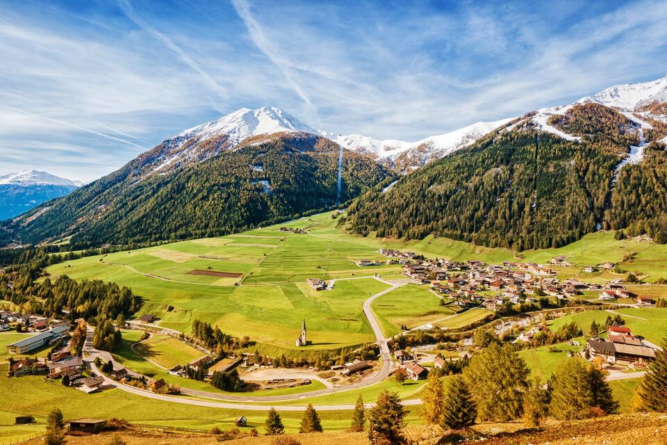 Un pueblo de montaña con picos nevados, campos verdes y un camino sinuoso.