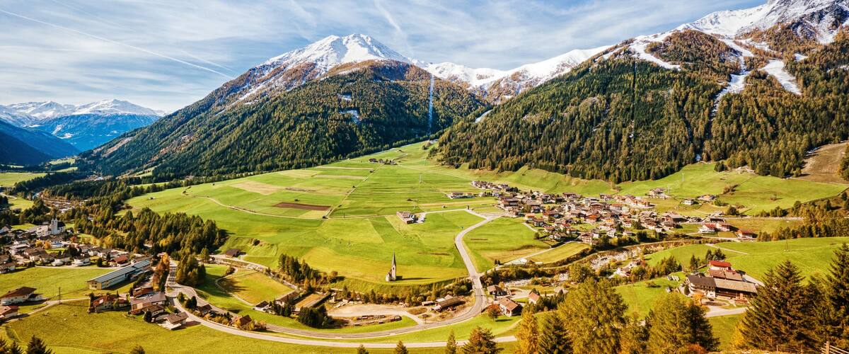A mountain village with snow-capped peaks, green fields, and a winding road.