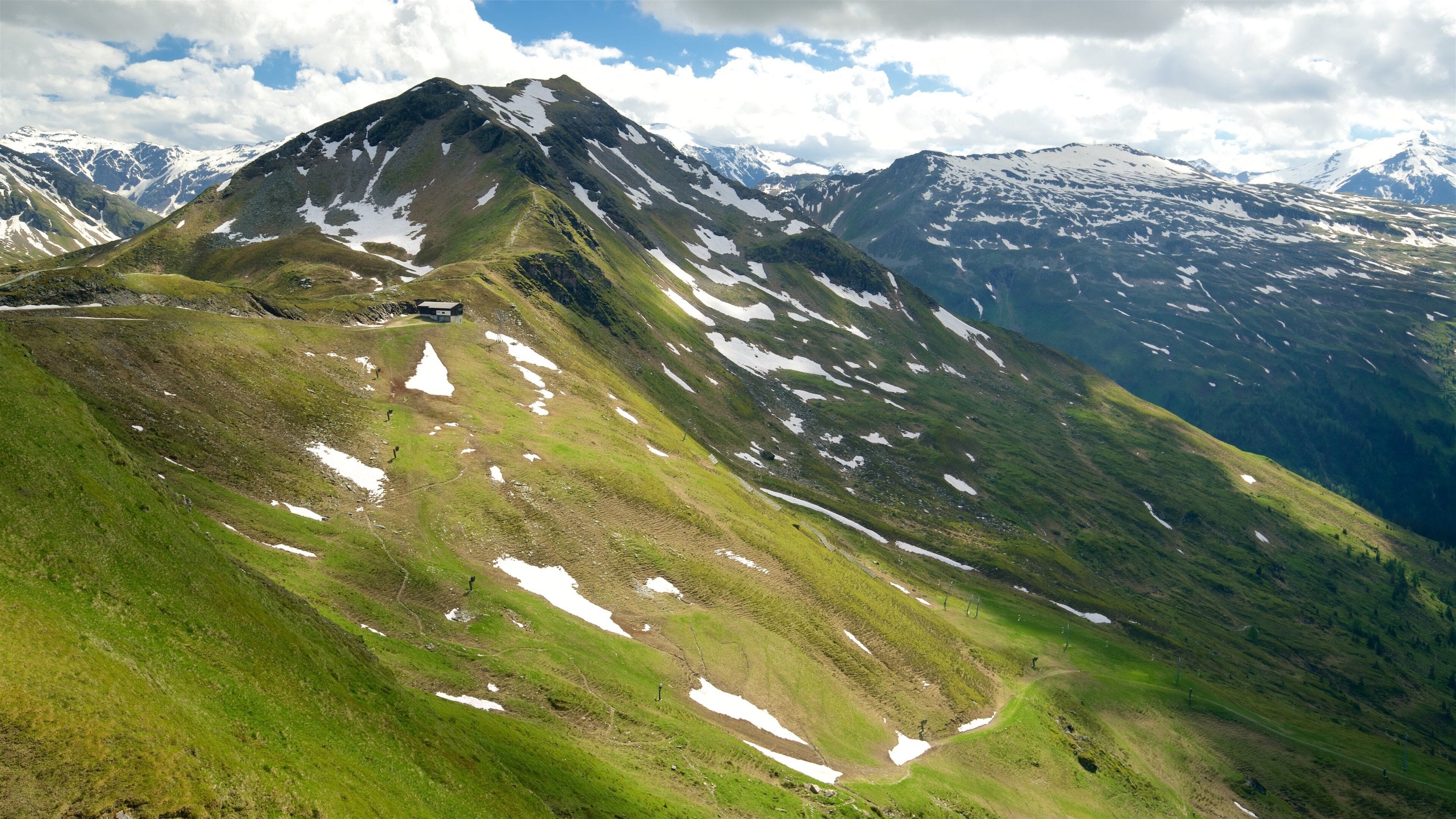 A mountain with patches of snow and green grass, a clear sky, and distant snow-capped peaks.