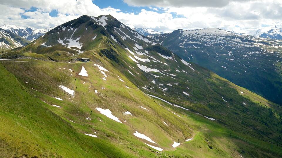 A mountain with patches of snow and green grass, a clear sky, and distant snow-capped peaks.