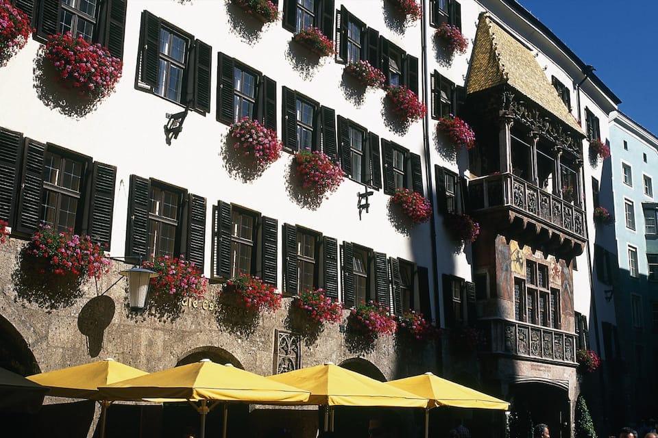 A white building with black shutters and flower boxes, yellow umbrellas, and a stone archway.