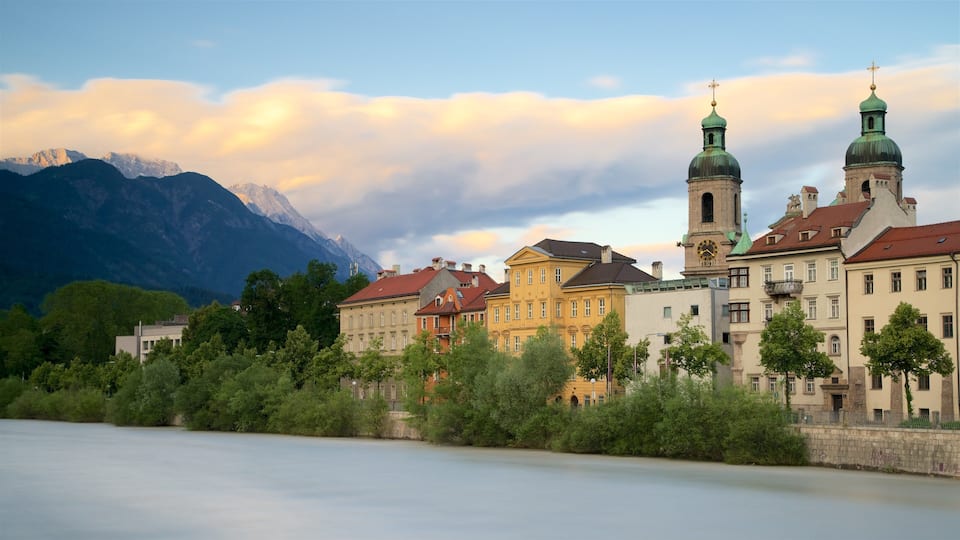 A historic town with colorful buildings, a river, and mountains in the background.