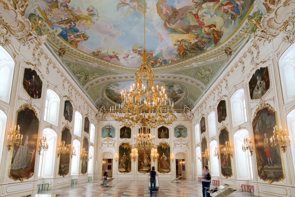An opulent room with a large chandelier, ornate ceiling frescoes, and classical paintings.