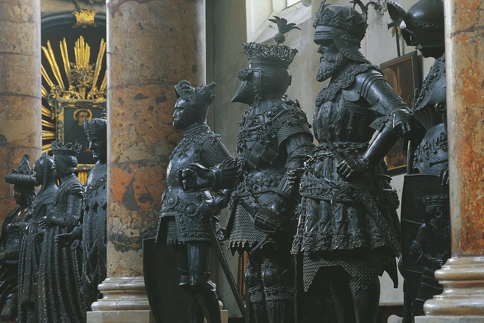 A group of statues in historical attire, set against a backdrop of classical architecture with columns and a golden emblem.