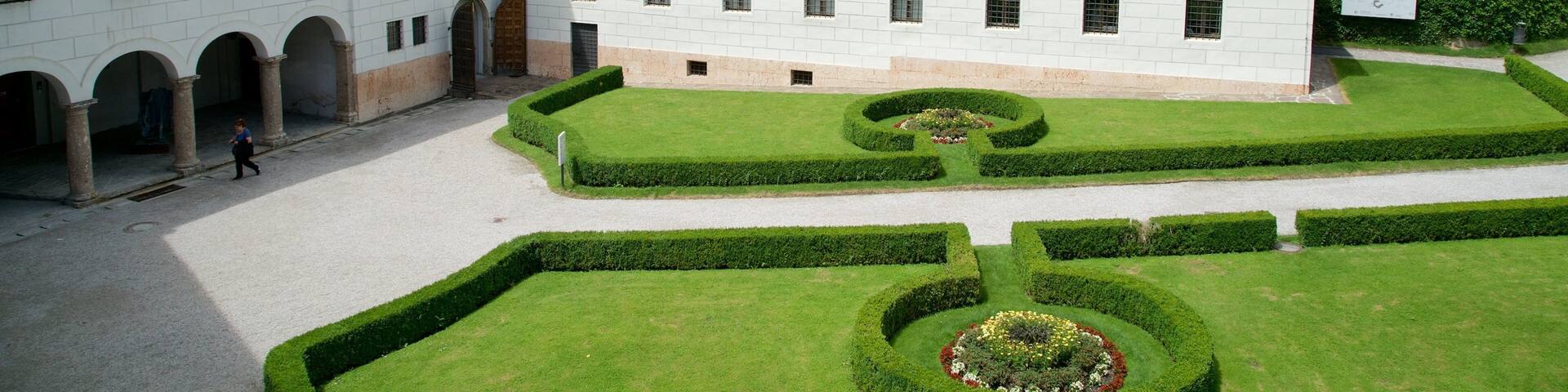 A historic building with a red-tiled roof, surrounded by well-manicured gardens and a paved courtyard.