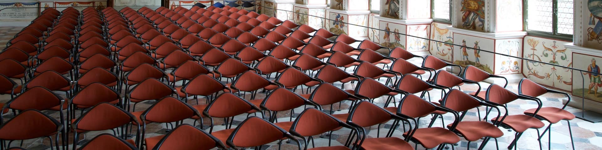 A grand hall with ornate ceilings, large windows, and rows of red chairs.