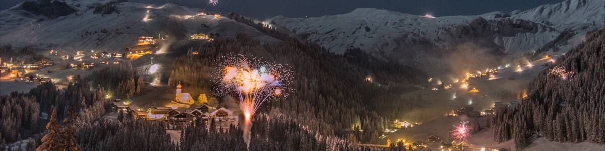 Ein schneebedecktes Gebirge bei Nacht mit einer Stadt im Tal und Feuerwerk im Vordergrund.