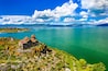 A historic church on a cliff overlooking a turquoise lake with mountains in the distance.