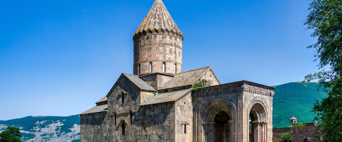 A historic stone church with a bell tower, surrounded by a grassy field and mountains in the background.
