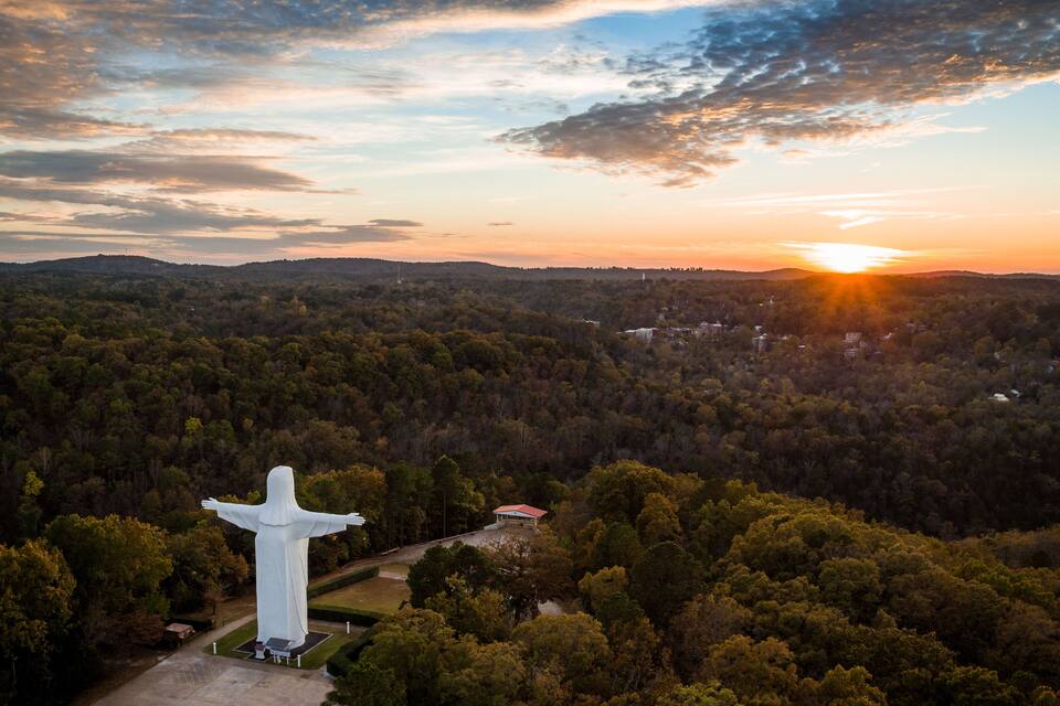 A large white statue with outstretched arms in a forested area with a sunset in the background.