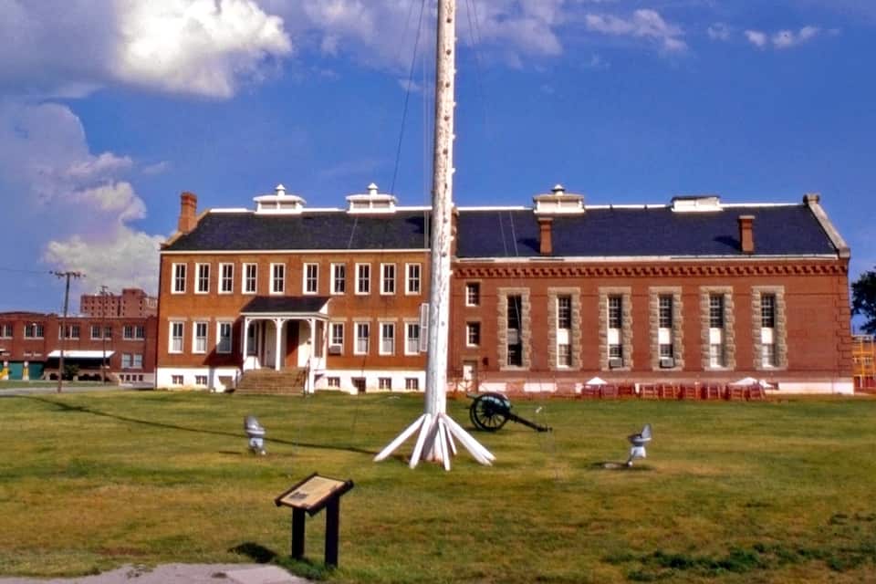 Un grand bâtiment en briques, doté d’une entrée centrale et d’un mât sur lequel flotte un drapeau, situé à l’avant.