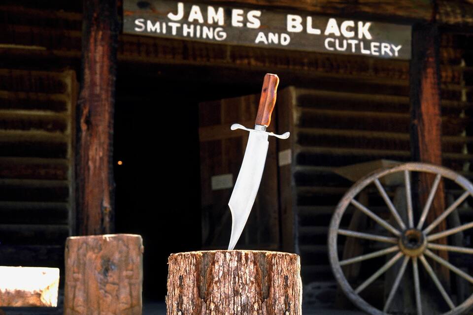 A wooden post with a knife stuck in it, in front of a building with a sign that reads "James Black Smithing and Cutlery".