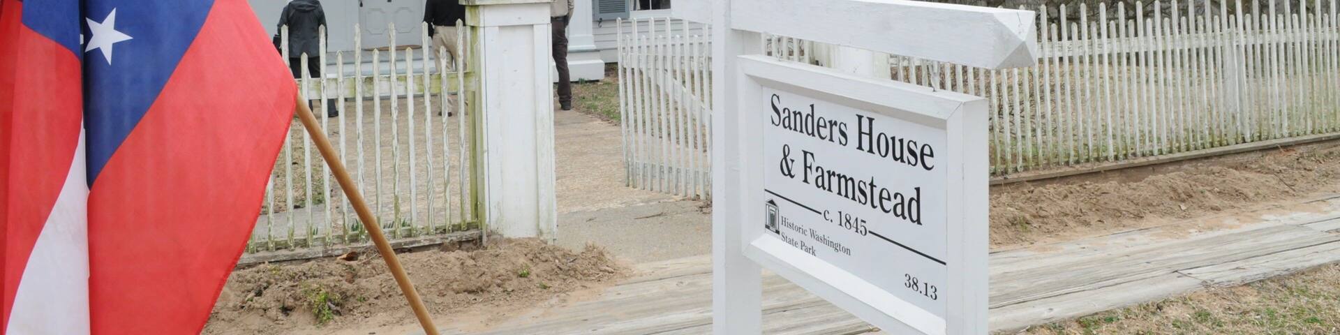 The exterior of the Sanders House & Farmstead, a historic building dating back to 1845, located at 38.13 on a street with a white picket fence and a flag.