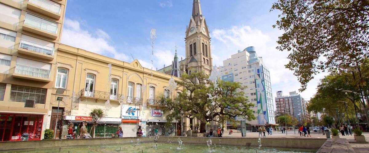 A fountain in a square with buildings, including a church tower, and a tree.