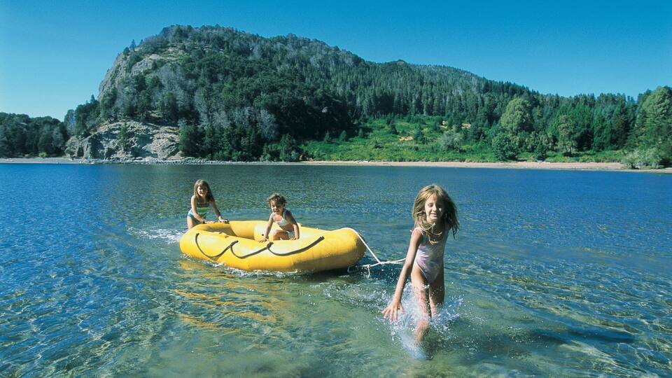 Three children on a yellow inflatable raft in a clear lake with a forested mountain in the background.