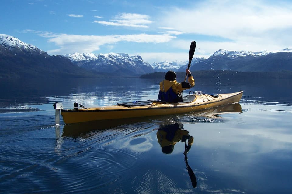 Una persona fa kayak su un lago calmo, con montagne innevate sullo sfondo.