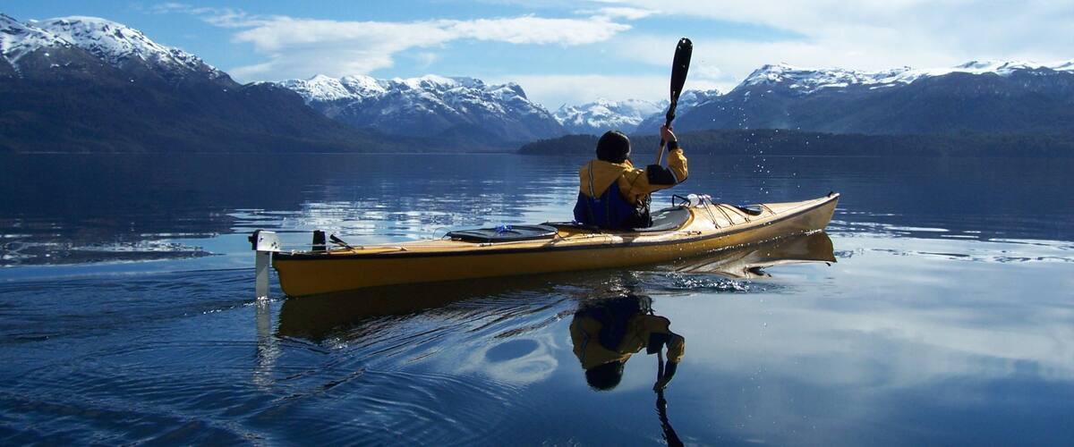 A person kayaking on a calm lake with snow-capped mountains in the background.