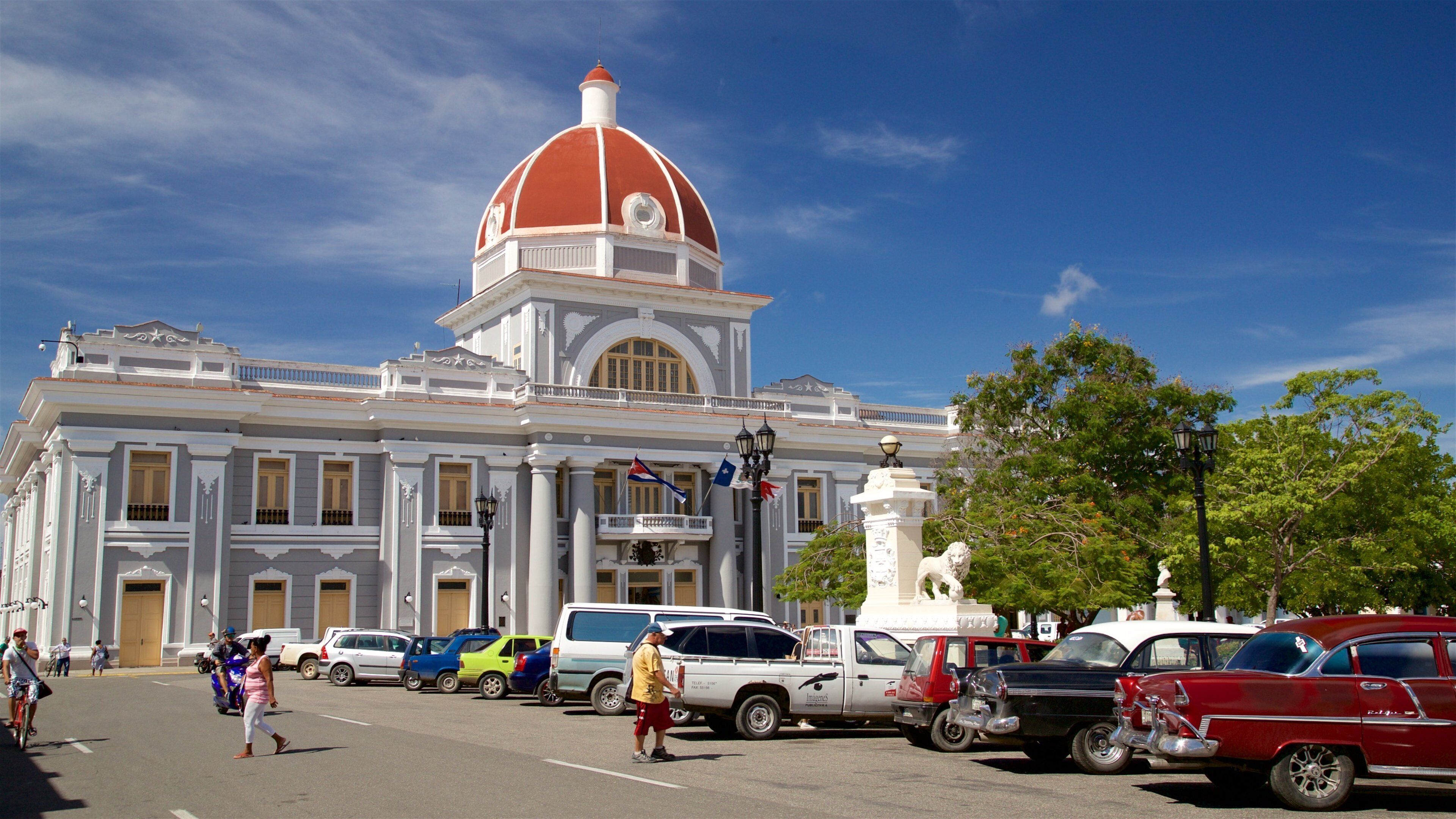 A historic building with a red dome and white columns, parked cars, and pedestrians.