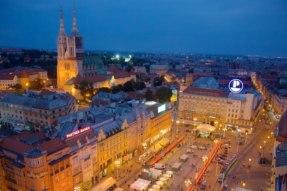 Une vue sur la ville au crépuscule, avec des bâtiments illuminés, un clocher d’église imposant et une place animée où se trouvent des vendeurs de rue.