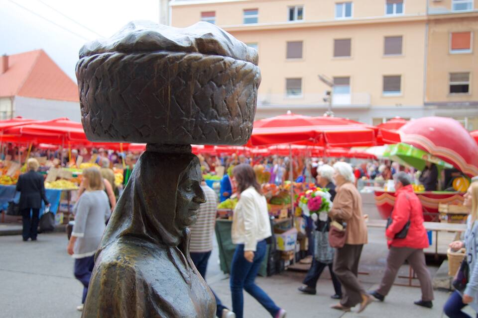 Un marché animé en plein air, avec des parasols rouges, des gens qui font leurs courses et une statue au premier plan.