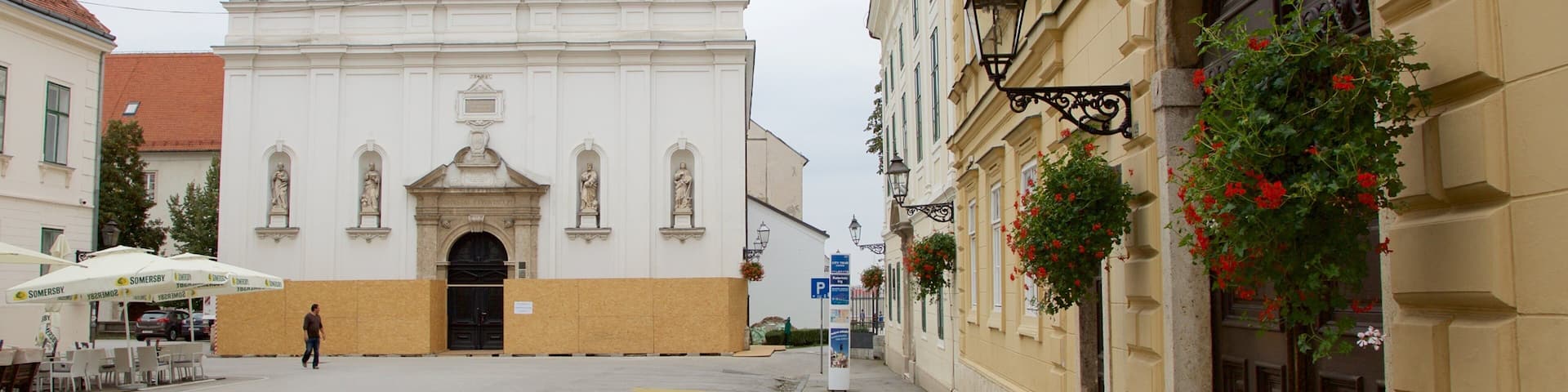A white church with a triangular roof, a blue flag with a coat of arms, and a building with a beige facade and red flowers.