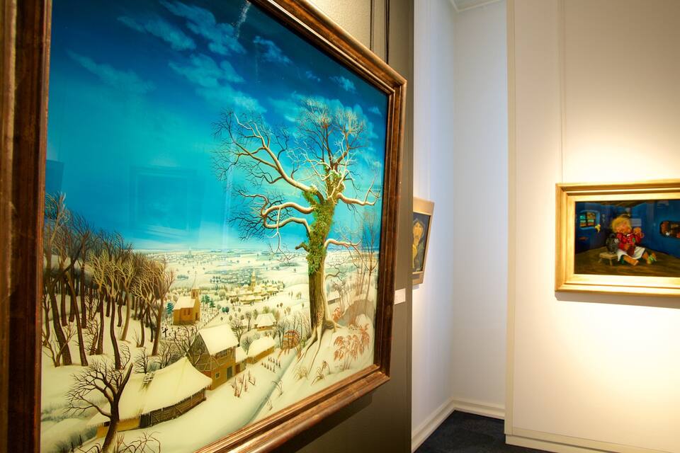 Un gran quadre de paisatge que mostra un poble cobert de neu amb un arbre destacat, i un quadre petit emmarcat que representa una persona a la dreta.