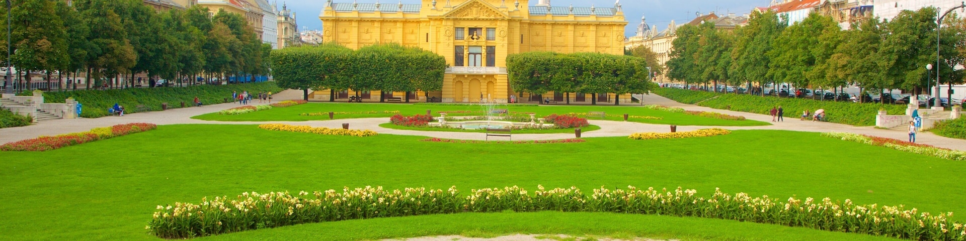 Ein Park mit einem zentralen Brunnen, Bänken und einem großen, gelben Gebäude mit Kuppel in der Ferne.