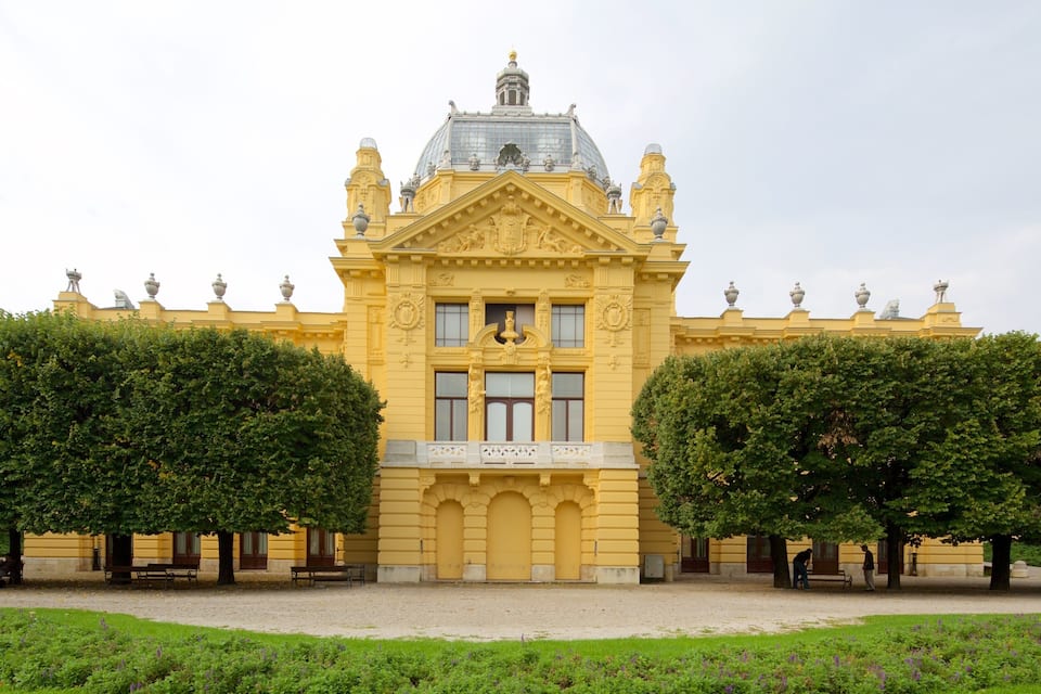 Un edifici gran i groc amb una cúpula i estàtues, envoltat d'arbres ben podats i un camí de grava.