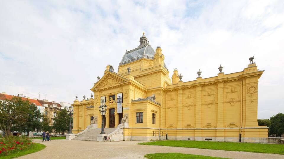 Un gran edificio amarillo con una cúpula y estatuas, rodeado de un parque verde y algunas personas caminando.