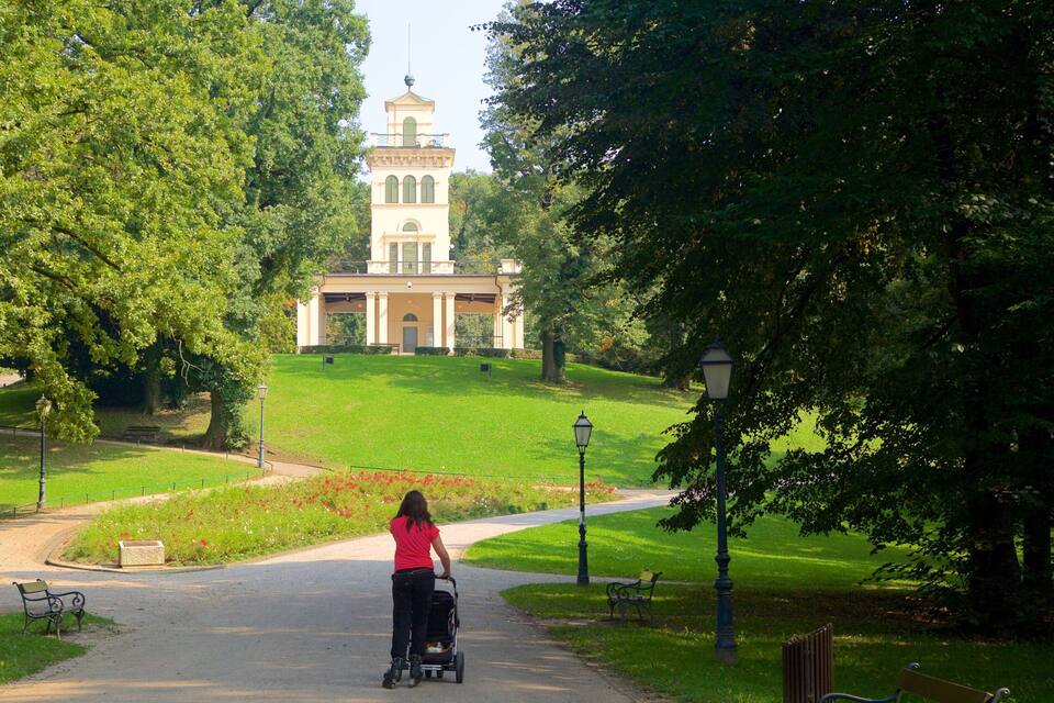 A woman pushing a stroller on a paved path in a park with a large building in the background.