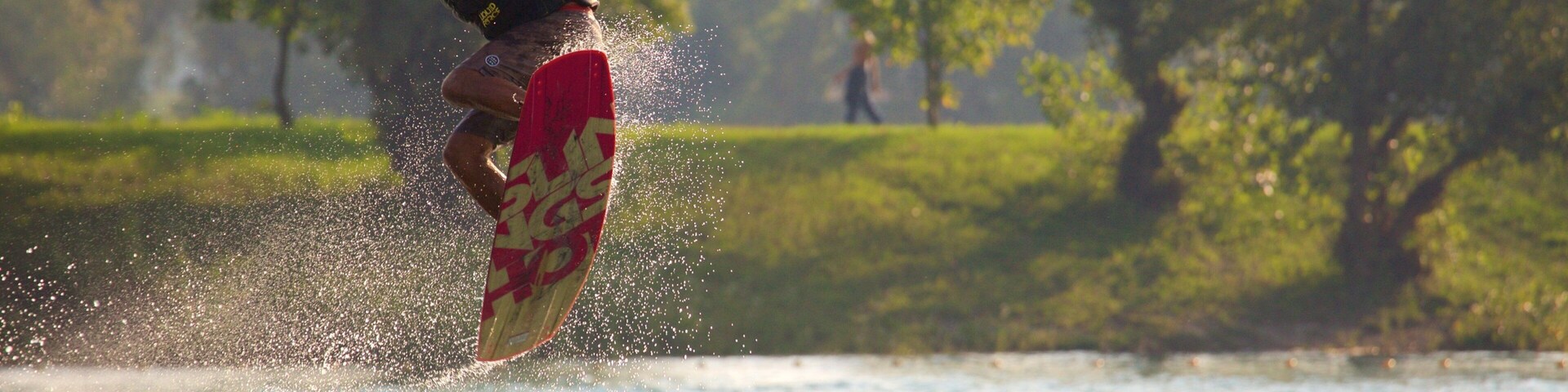 Uma pessoa praticando wakeboard sobre a água, com respingos, uma prancha vermelha e árvores ao fundo.