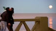 Una pareja besándose en un puente, con la luna de fondo.