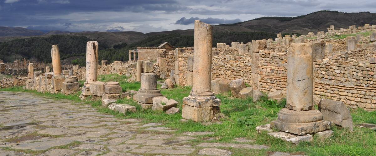 Ancient ruins with stone columns and walls, set against a backdrop of hills and a cloudy sky.