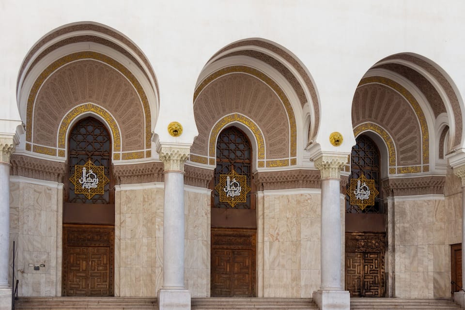 Fachada de un edificio con tres grandes puertas de madera, cada una enmarcada por un arco adornado con intrincados azulejos y un emblema dorado.