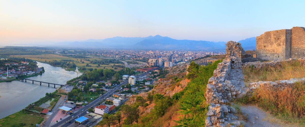 A historic stone fortress overlooking a city with a river and mountains in the background.