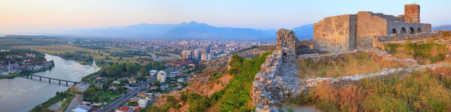 A historic stone fortress overlooking a city with a river and mountains in the background.