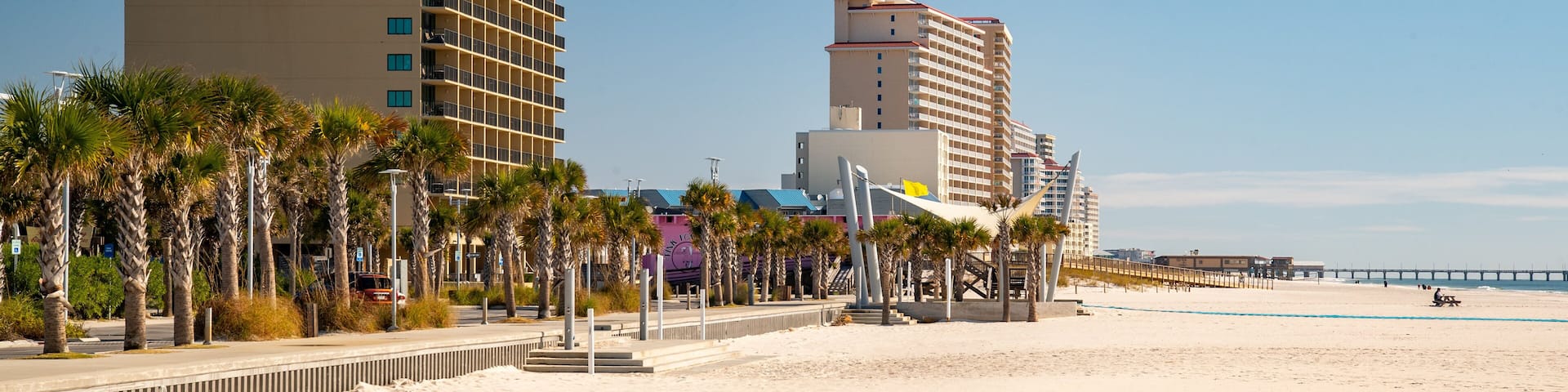 A beach with palm trees, a boardwalk, and buildings including a hotel.