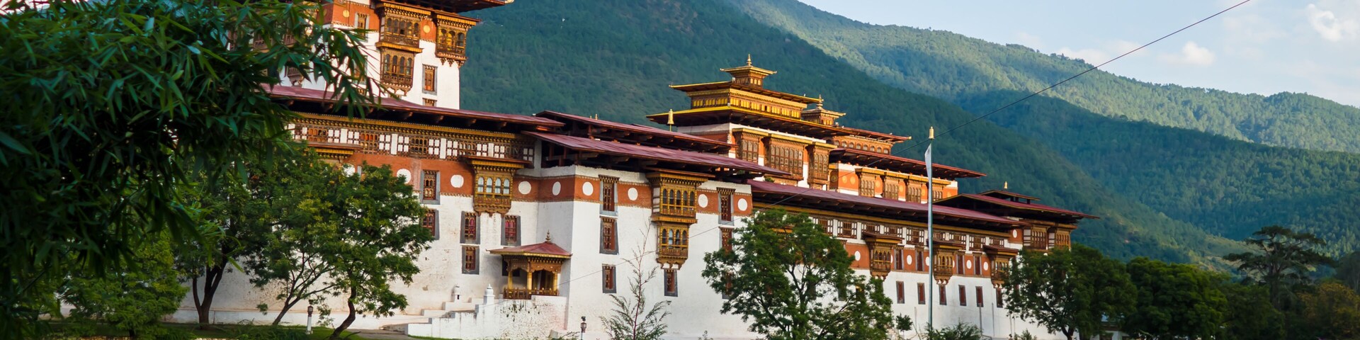 A traditional building with multiple tiers and red roofs, situated by a river with mountains in the background.