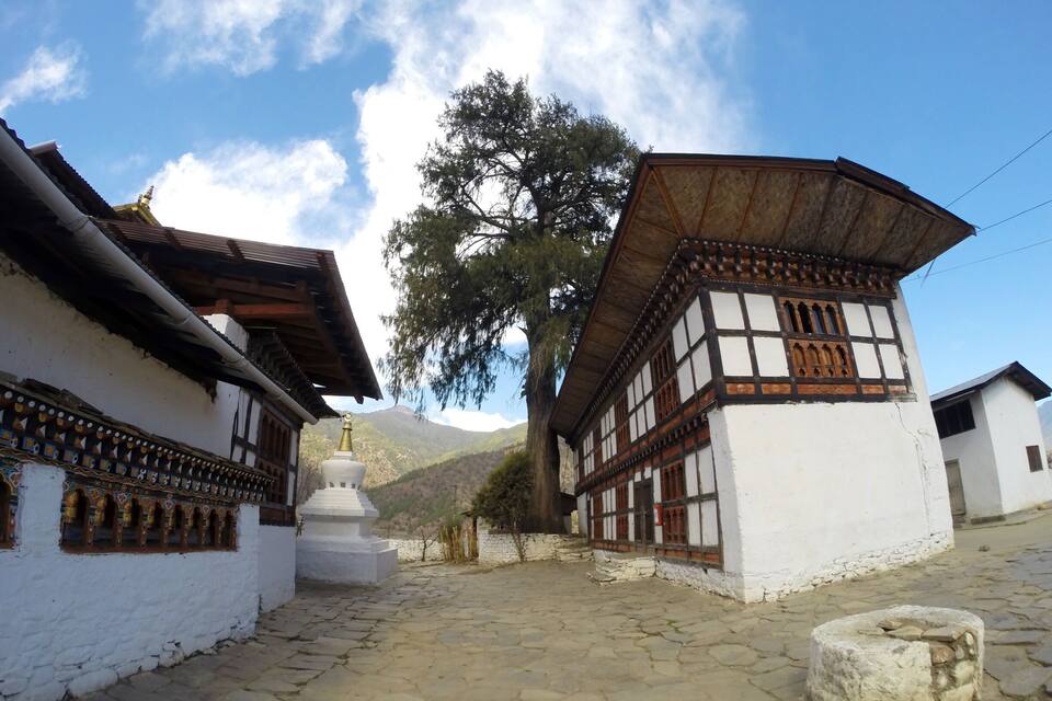 Un edificio tradicional con fachada blanca y detalles de madera, un patio empedrado de piedra y una montaña al fondo.
