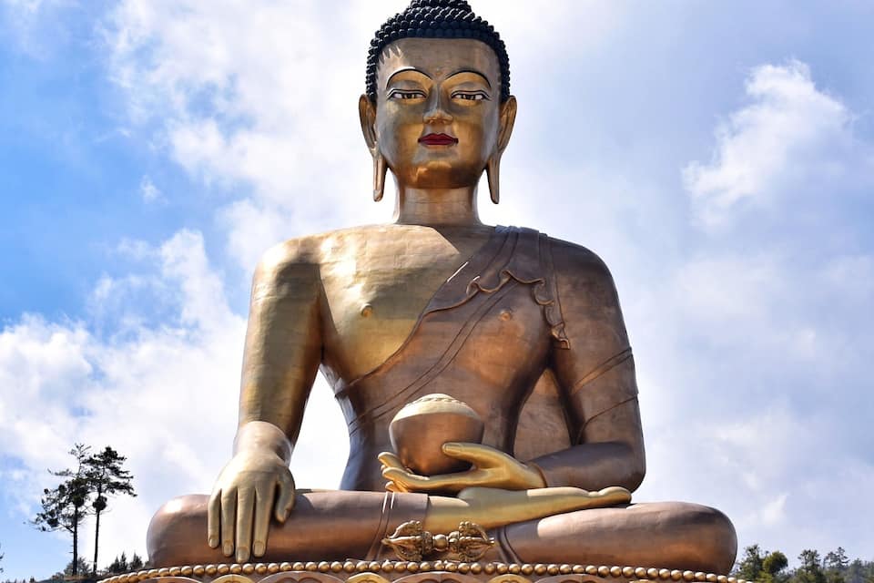 A large seated Buddha statue with intricate carvings on the base, set against a clear blue sky with scattered clouds.