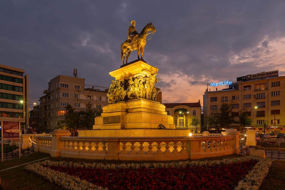 A golden statue of a figure on horseback, surrounded by a flower bed, with modern buildings in the background.