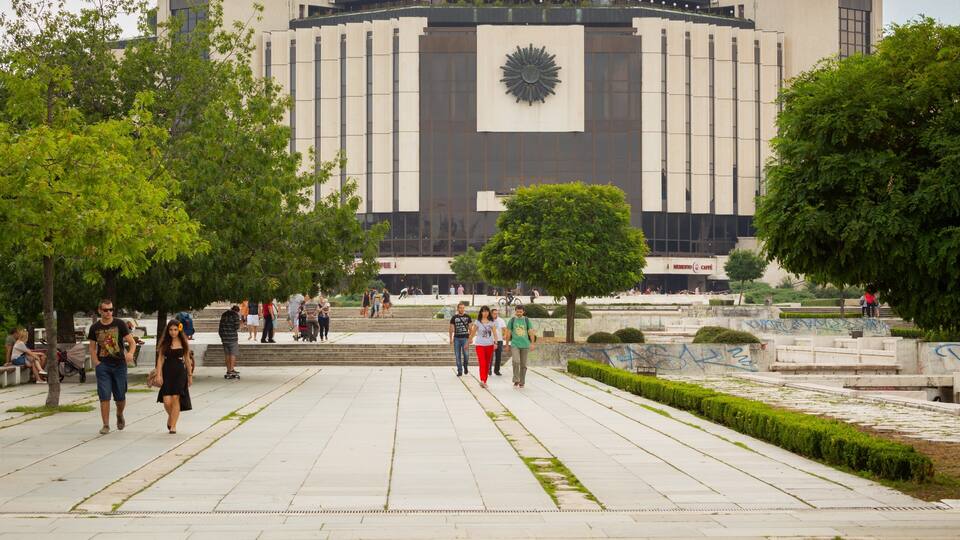 Une place publique avec un grand bâtiment dont la façade présente un motif rappelant un soleil, entourée d’arbres et animée par la présence de passants.