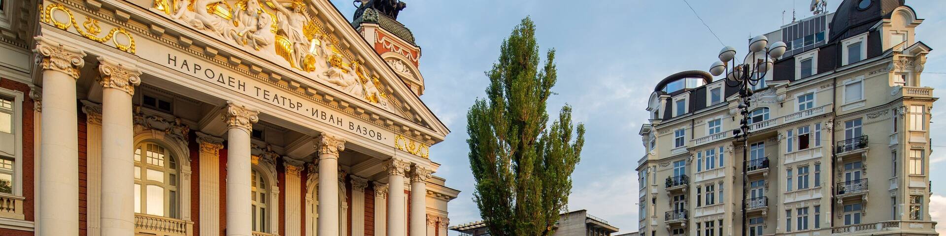 Un gran teatro con columnas y estatuas clásicas, rodeado por una plaza con pavimento estampado y edificios modernos.