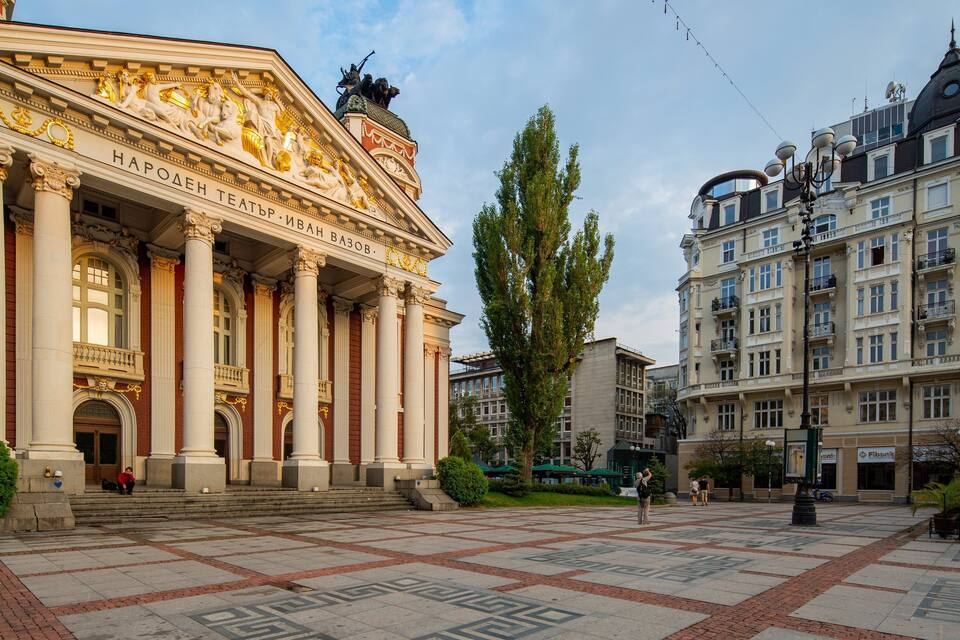Un gran teatro con columnas y estatuas clásicas, rodeado por una plaza con pavimento estampado y edificios modernos.